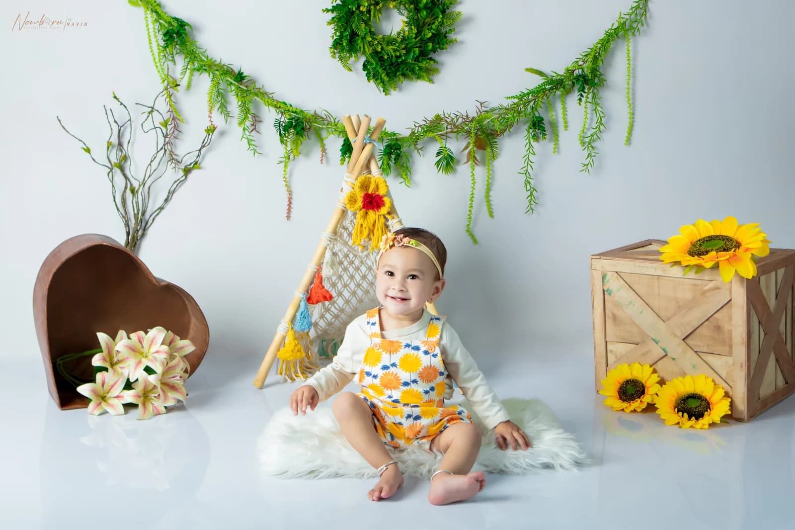 Smiling baby girl in a sunflower-print outfit sitting in a sunflower-themed boho photography set with a mini macrame teepee, greenery garland and a wooden crate.