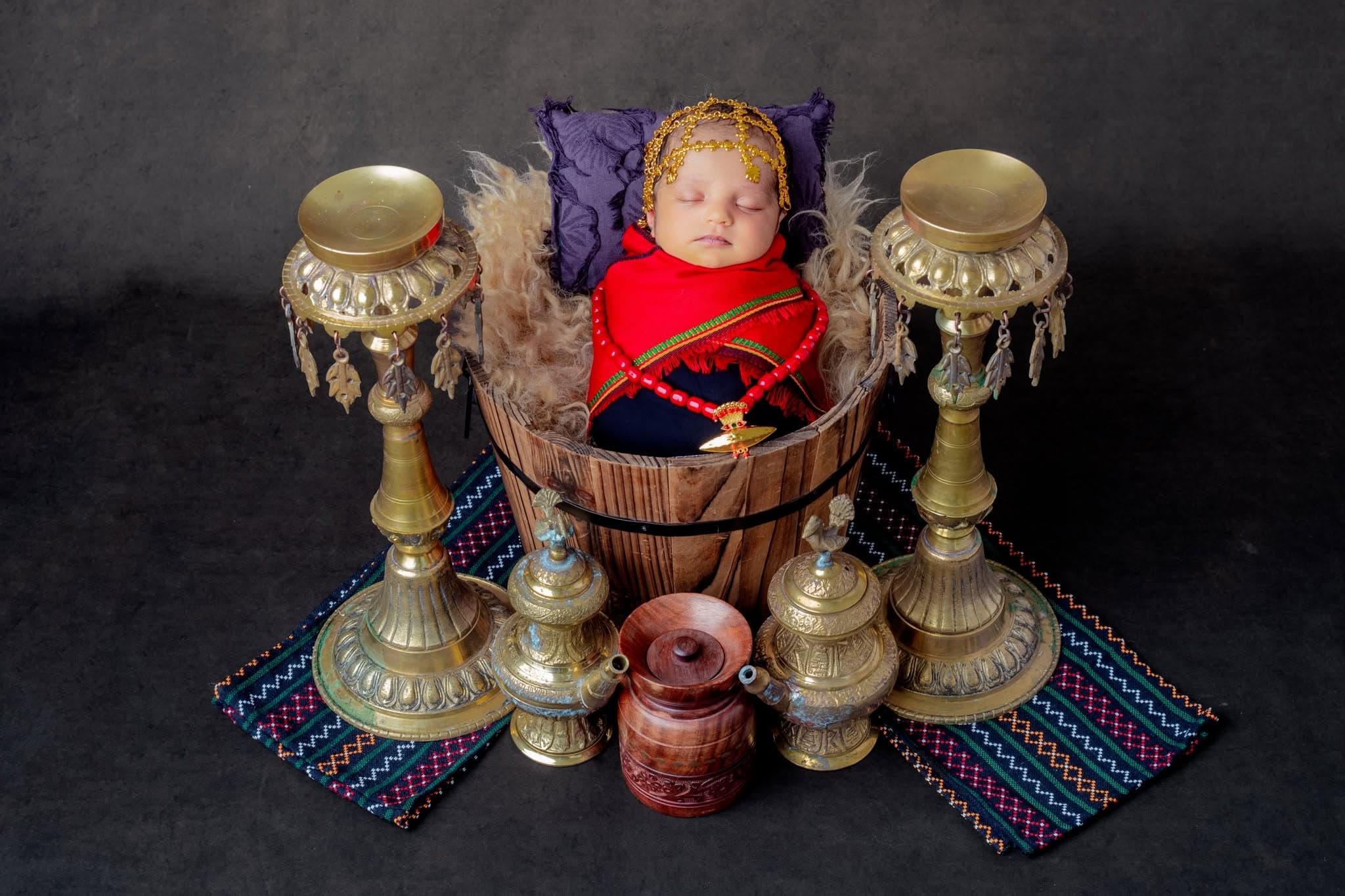 A sleeping newborn dressed in traditional Nepali headwear and a red wrap, styled in a wooden bucket surrounded by brass vessels and hand-woven textiles.