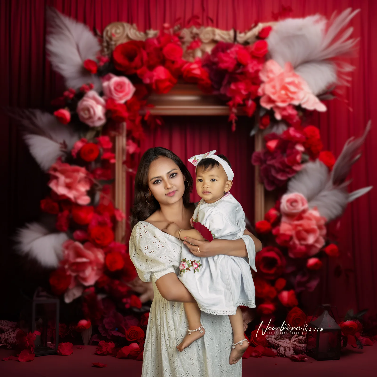 Mother in a white dress holding her one-year-old, dressed in white, in front of a red and pink floral photo frame with feathers.