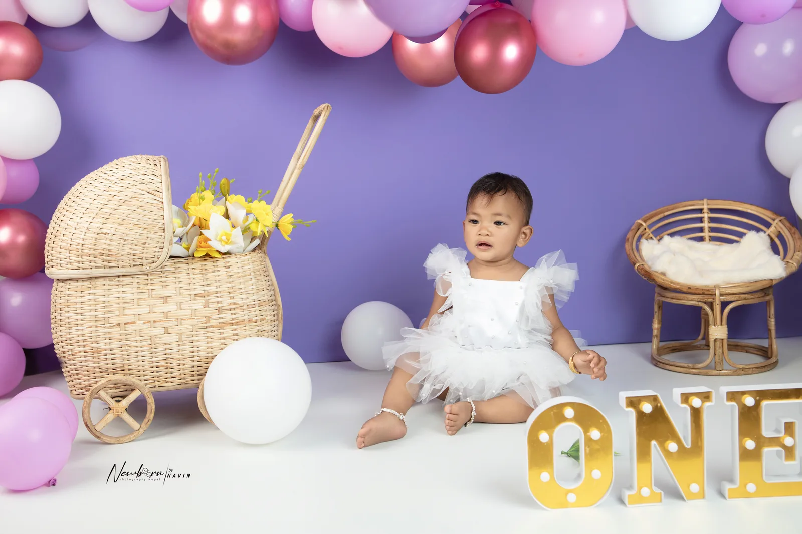 One-year-old girl in a white ruffled dress sitting beside a wicker pram and a fur-lined rattan chair, pastel balloon garland behind her.