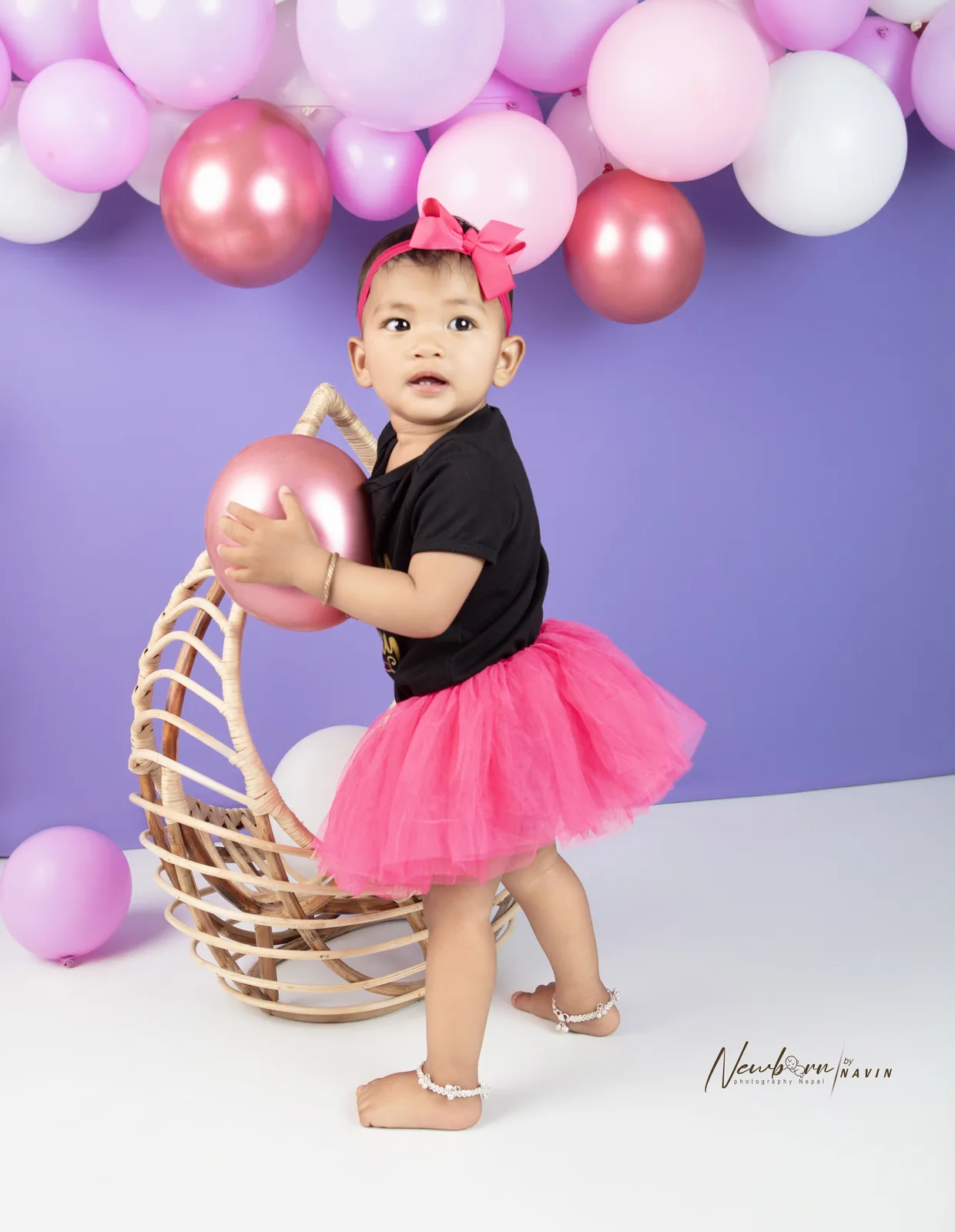 One-year-old girl in a black top and pink tutu holding a pink balloon beside a rattan hanging chair, purple balloon garland behind her.