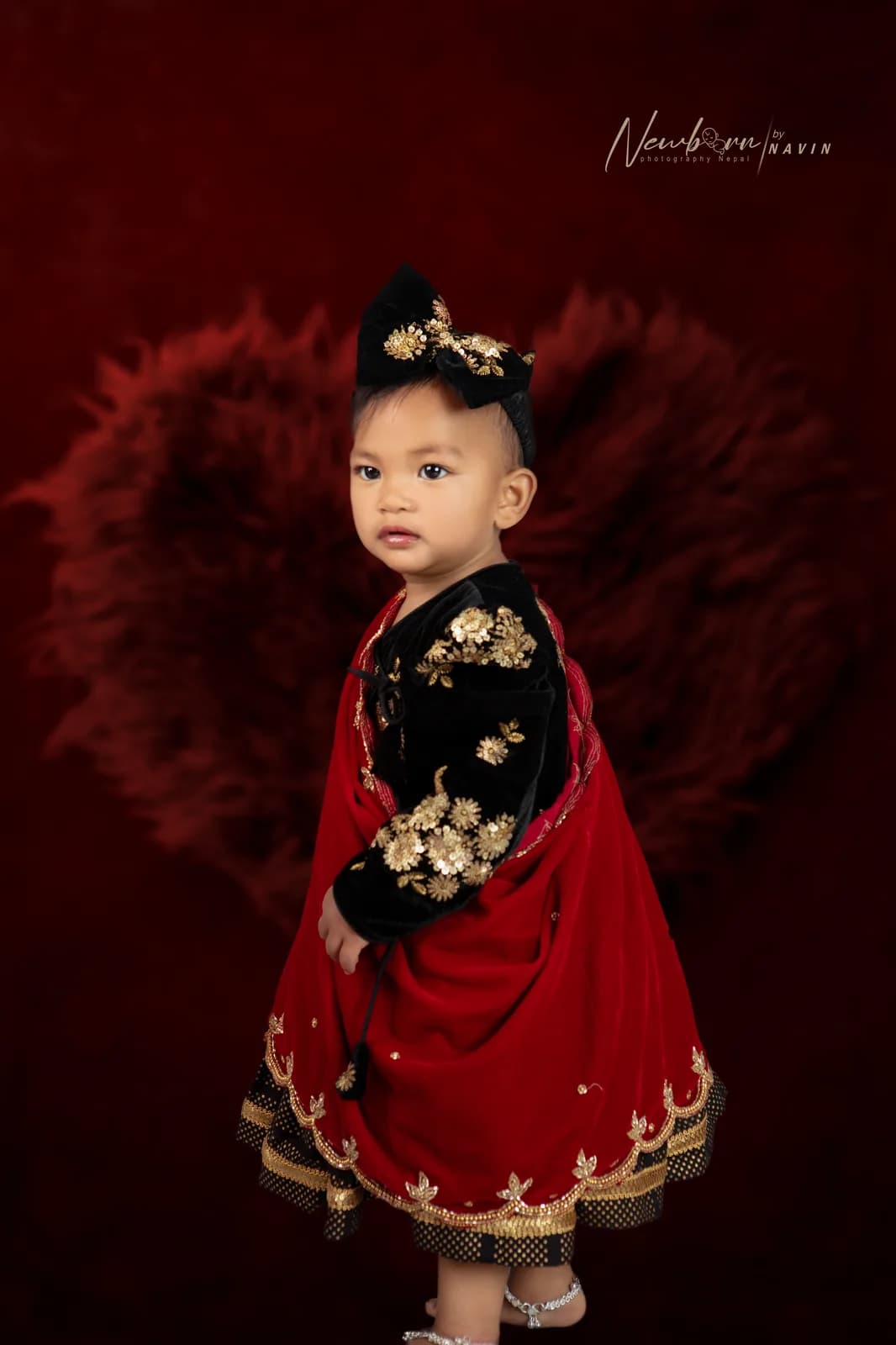 Toddler in traditional black and red Nepali dress with gold embroidery, photographed against a dark red feathered backdrop.