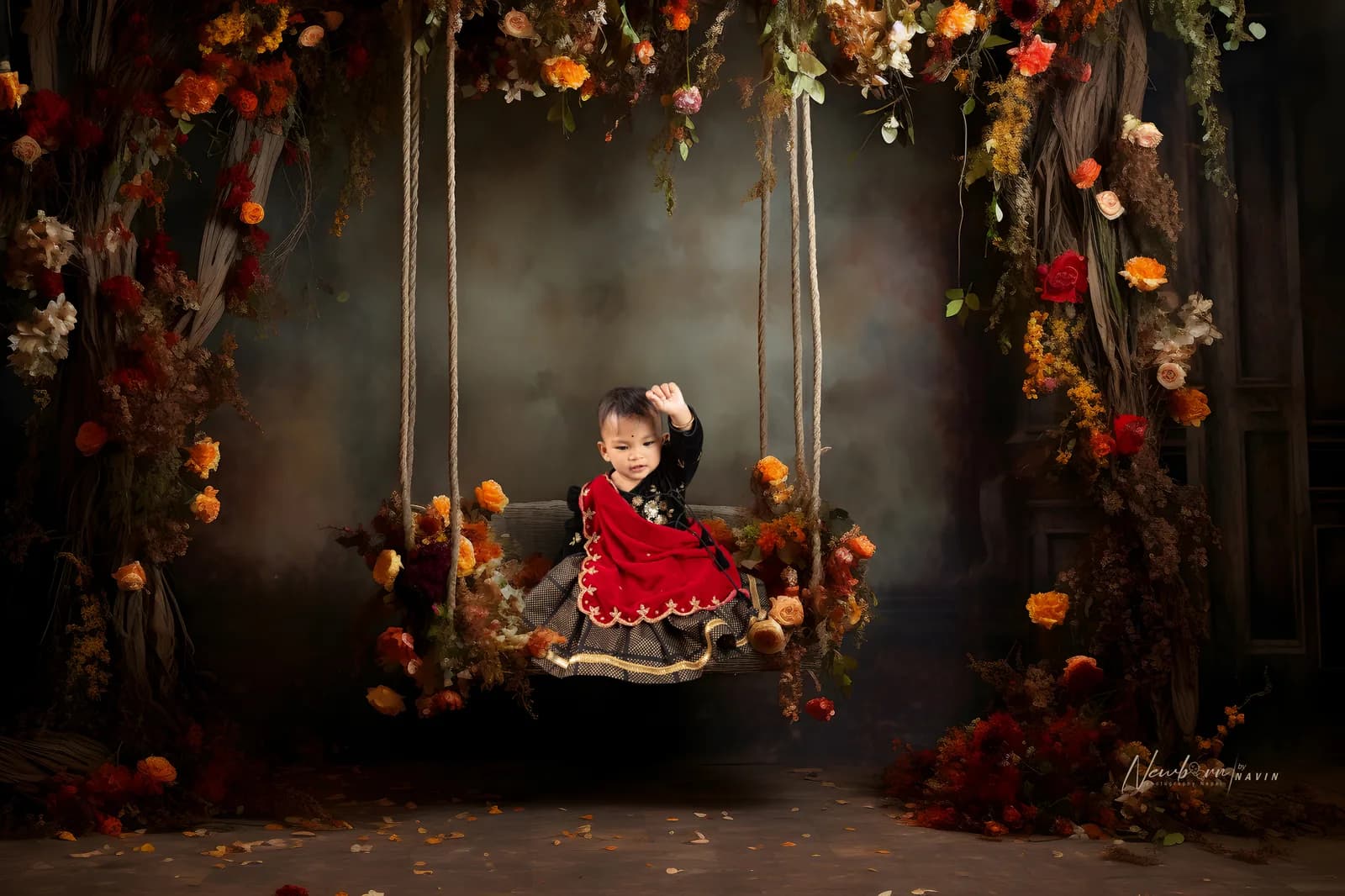 Toddler in traditional red and black Nepali dress, seated on a decorated swing surrounded by autumnal flowers.
