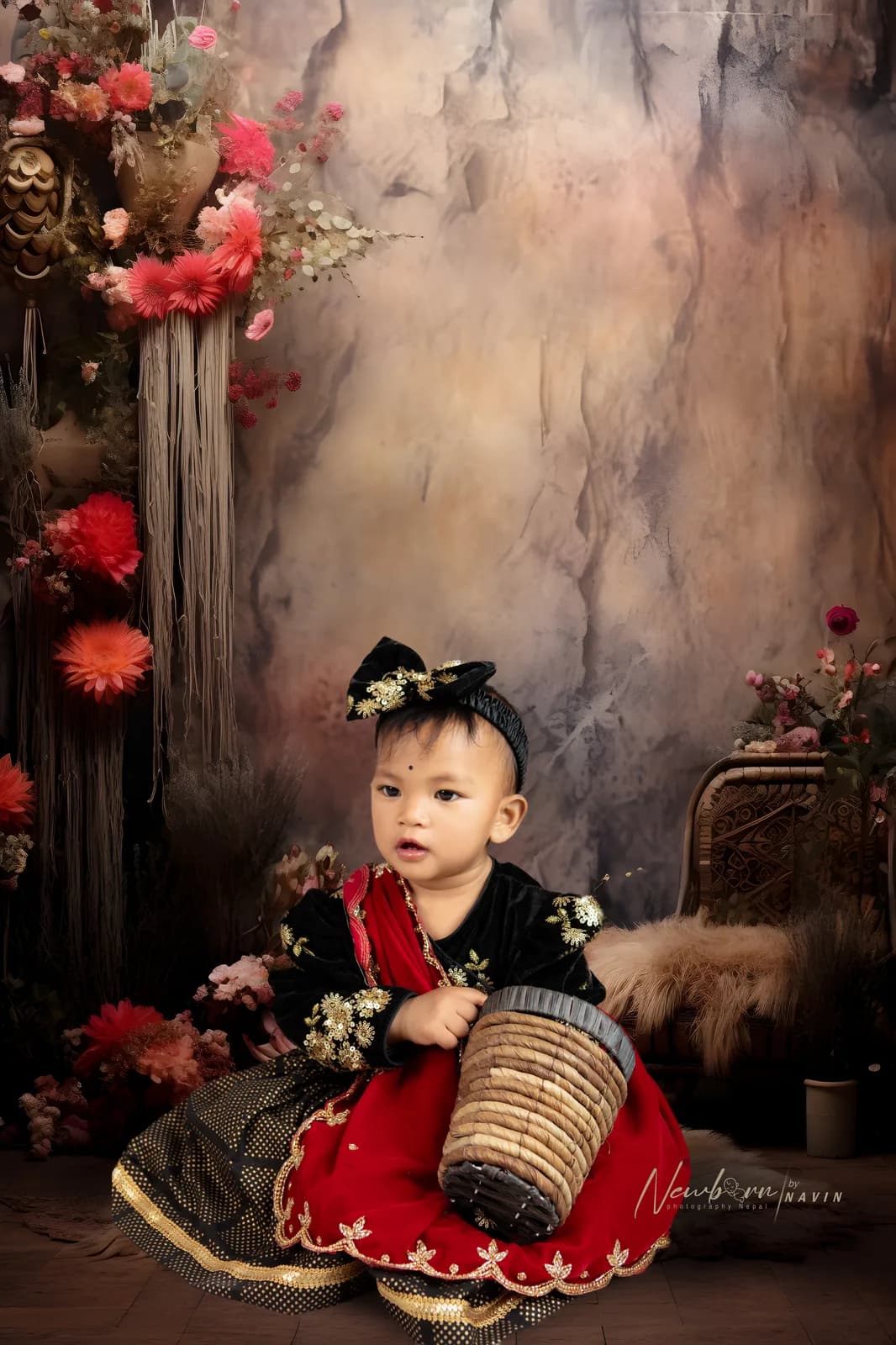 Toddler in traditional black and red Nepali dress holding a woven basket, seated beside floral décor and a fur-draped chair.