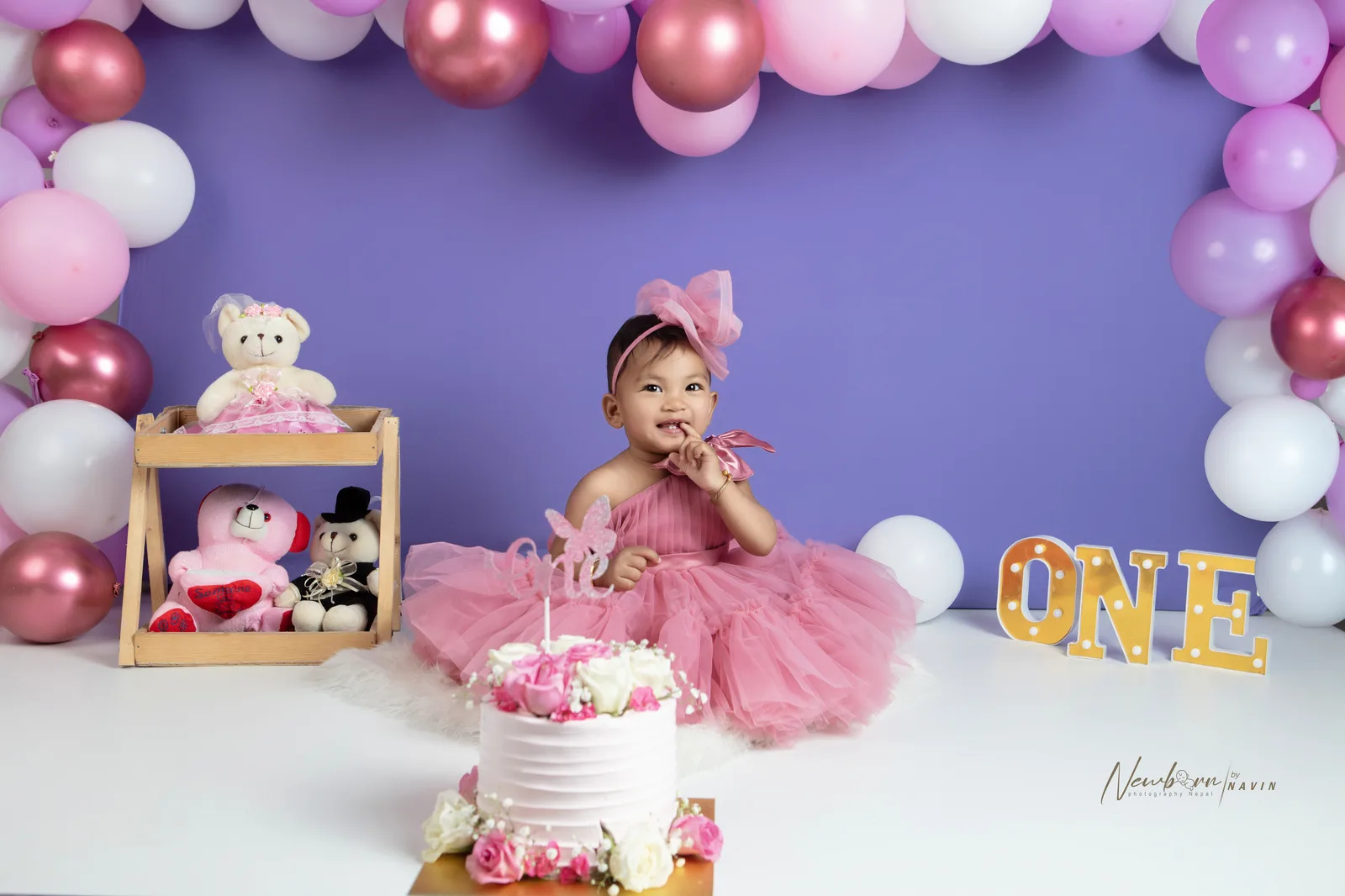 One-year-old girl in a pink tulle dress sitting beside her smash cake, finger raised, styled with a pink and purple balloon garland and a 'ONE' sign.