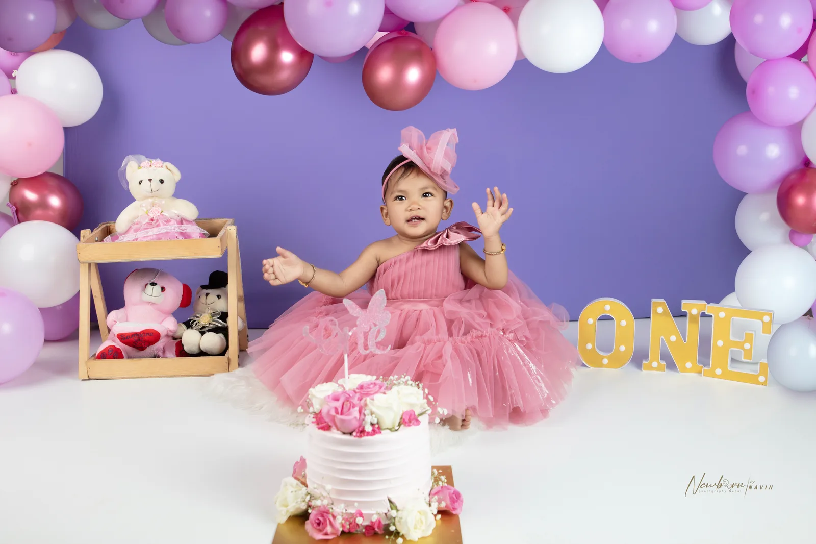 One-year-old girl in a pink dress with cake frosting on her face and hand raised, sitting beside her smashed cake.