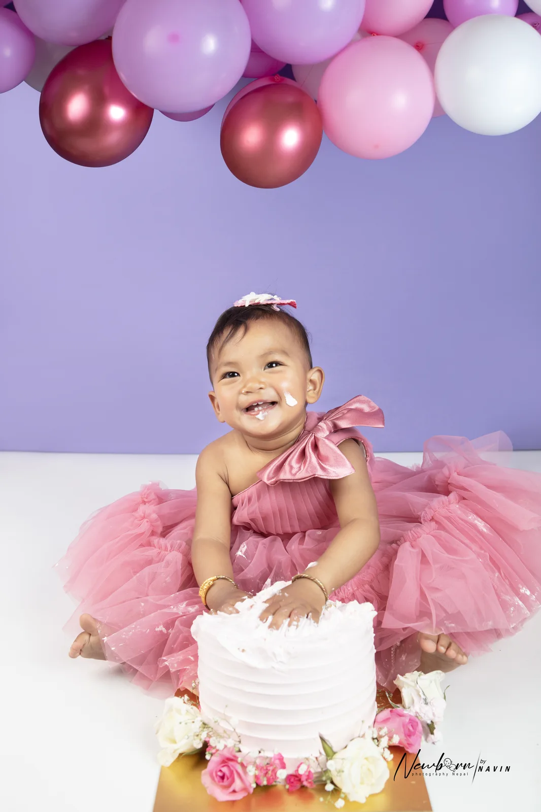 One-year-old girl in a pink dress with frosting on her cheek, smiling and touching her cake, purple and pink balloon garland behind her.