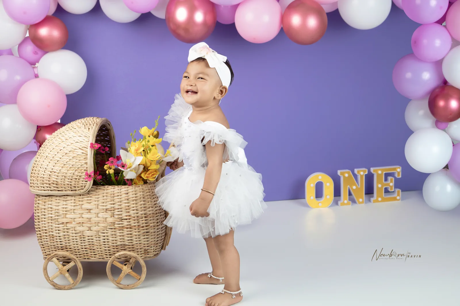 One-year-old girl in a white ruffled outfit standing beside a wicker pram styled with flowers, pink and purple balloon garland behind her.