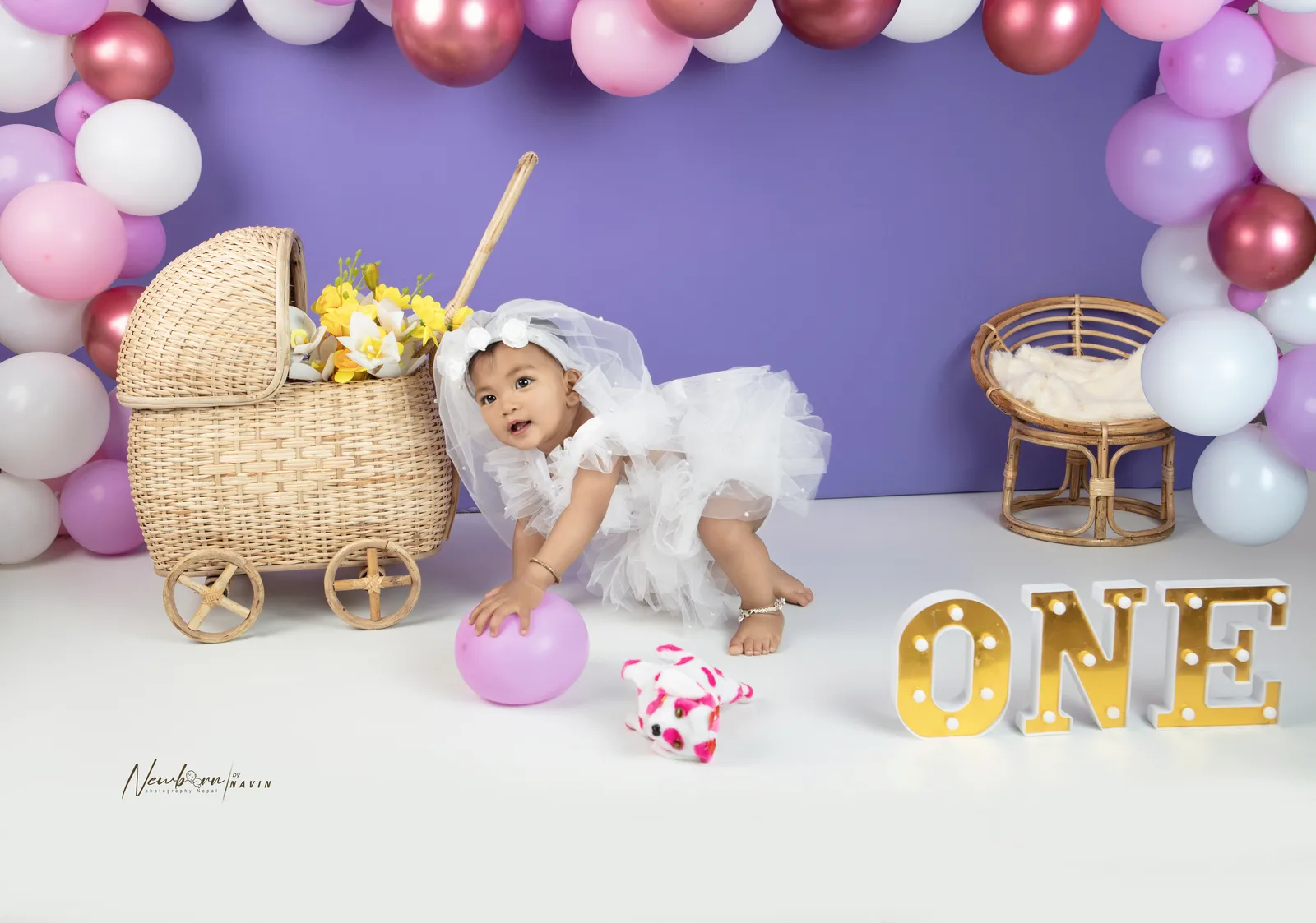 One-year-old girl in a white ruffled outfit and veil crawling toward a small pink ball, beside a wicker pram, balloon garland behind her.