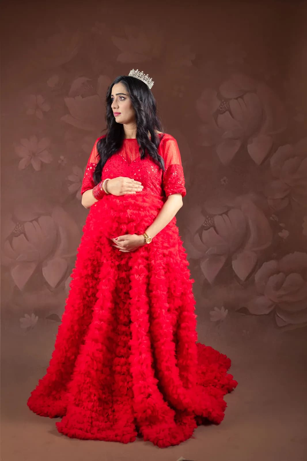 Pregnant woman in a red ruffled gown and a crystal crown, standing with one hand on her bump against a floral brown backdrop.