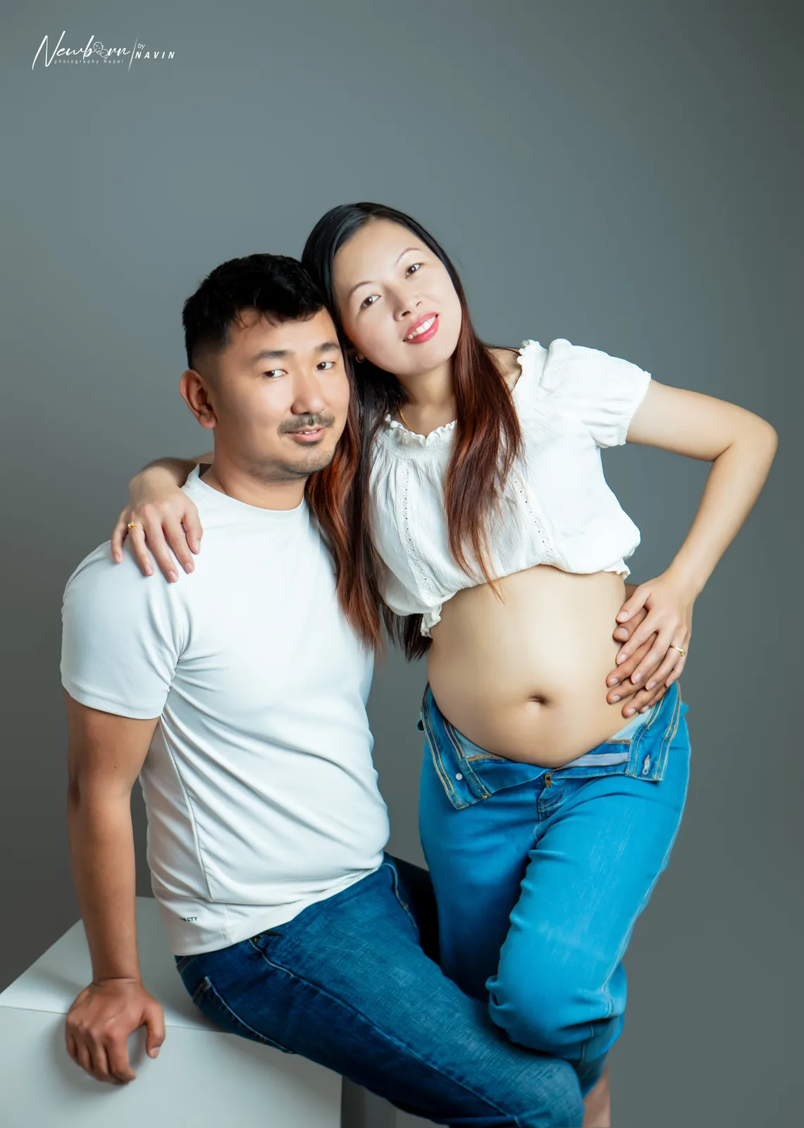 Expecting parents smiling together, the mother's hand on her bare bump, in a casual indoor setting.