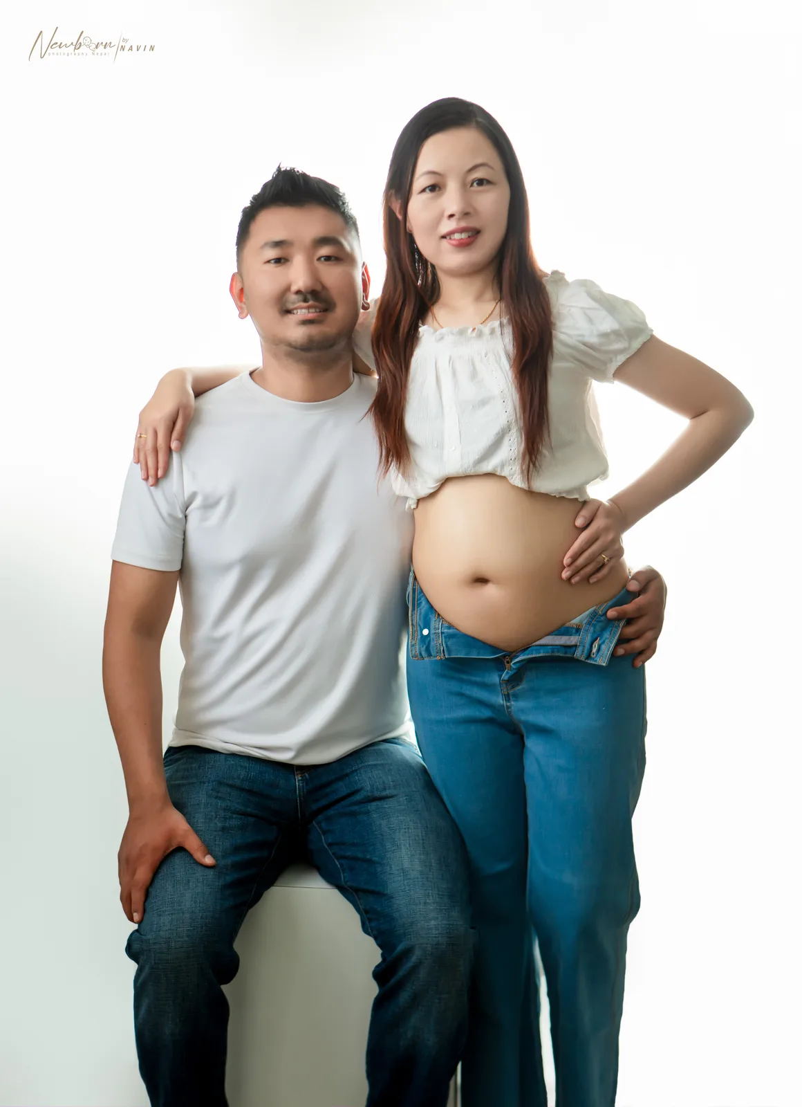 Expecting parents posing together, the mother resting her hand on her bare bump, against a plain backdrop.