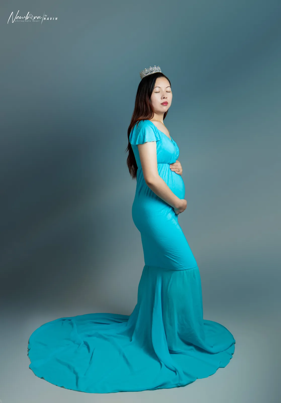 Pregnant woman in a teal gown with a crystal crown, one hand on her bump, in a three-quarter length side profile portrait.