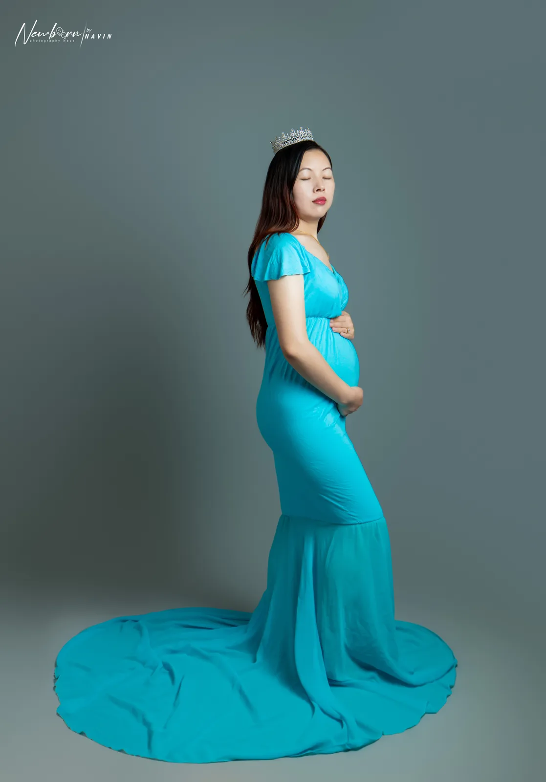 Pregnant woman in a teal gown with a crystal crown, one hand on her bump, in a full-length side profile portrait with her dress train sweeping across the floor.