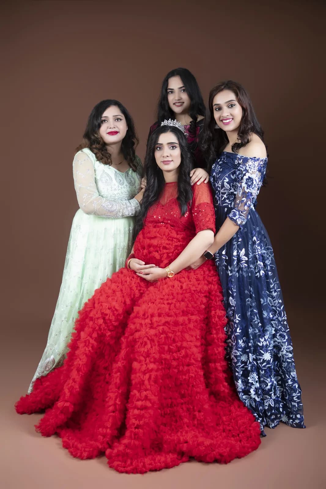 Four women in formal gowns posing together, with a pregnant woman in a red gown seated at the center, hands on her bump.