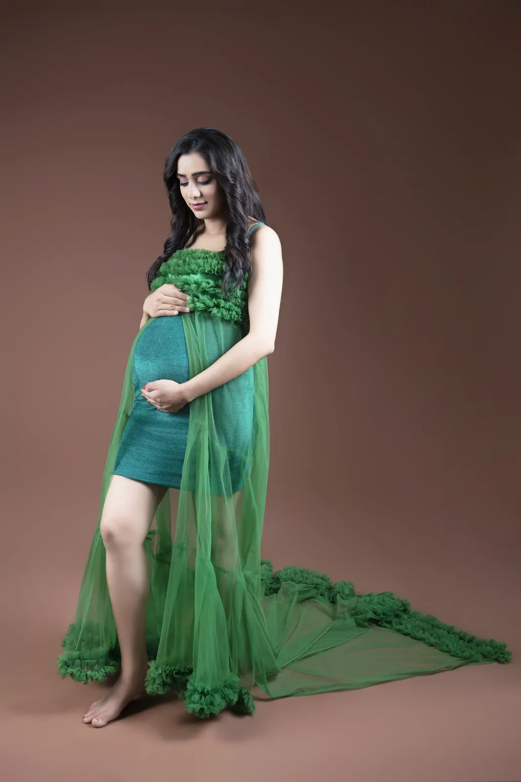Pregnant woman in a green off-shoulder dress with a sheer train, standing with one hand on her bump against a plain backdrop.