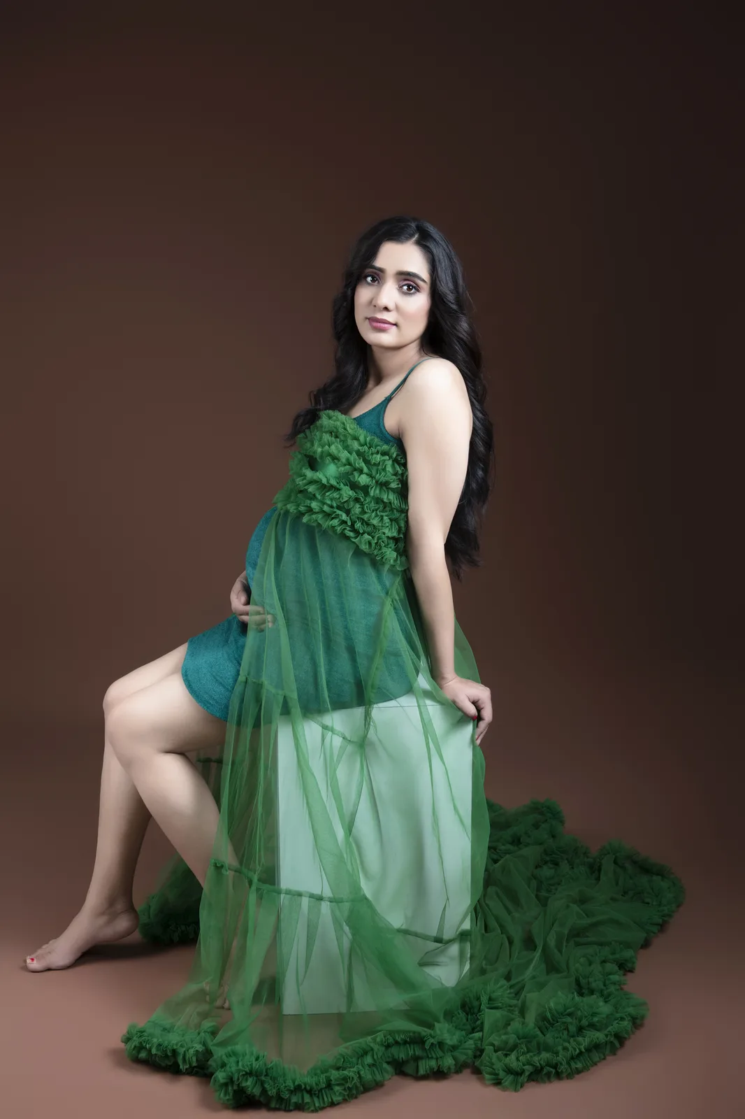 Pregnant woman in a green dress seated on a pedestal, one hand on her bump, sheer green fabric trailing across the floor.