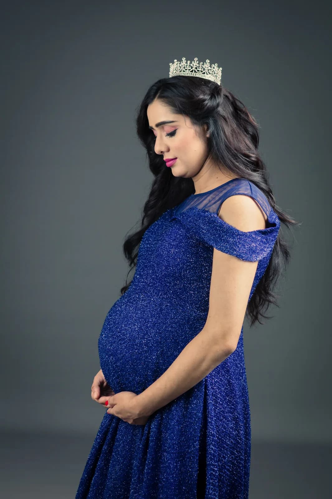 Pregnant woman in a flowing pink gown with fabric caught in motion, one hand on her bump, against a grey backdrop.