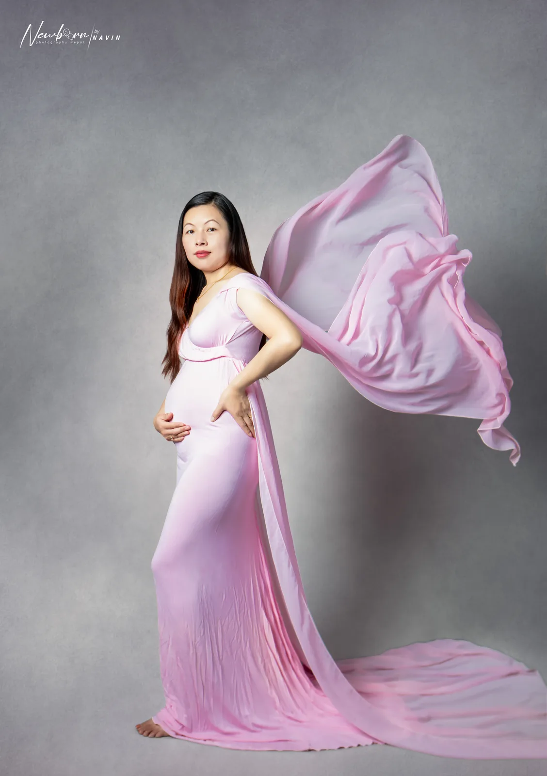 Pregnant woman in a flowing pink gown, one hand on her bump and one on her hip, against a grey backdrop.