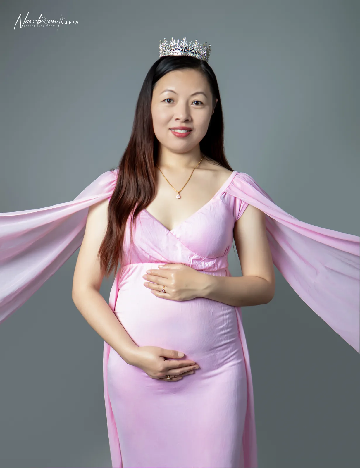 Pregnant woman in a pink gown with flowing sleeves, both hands on her bump, against a grey backdrop.