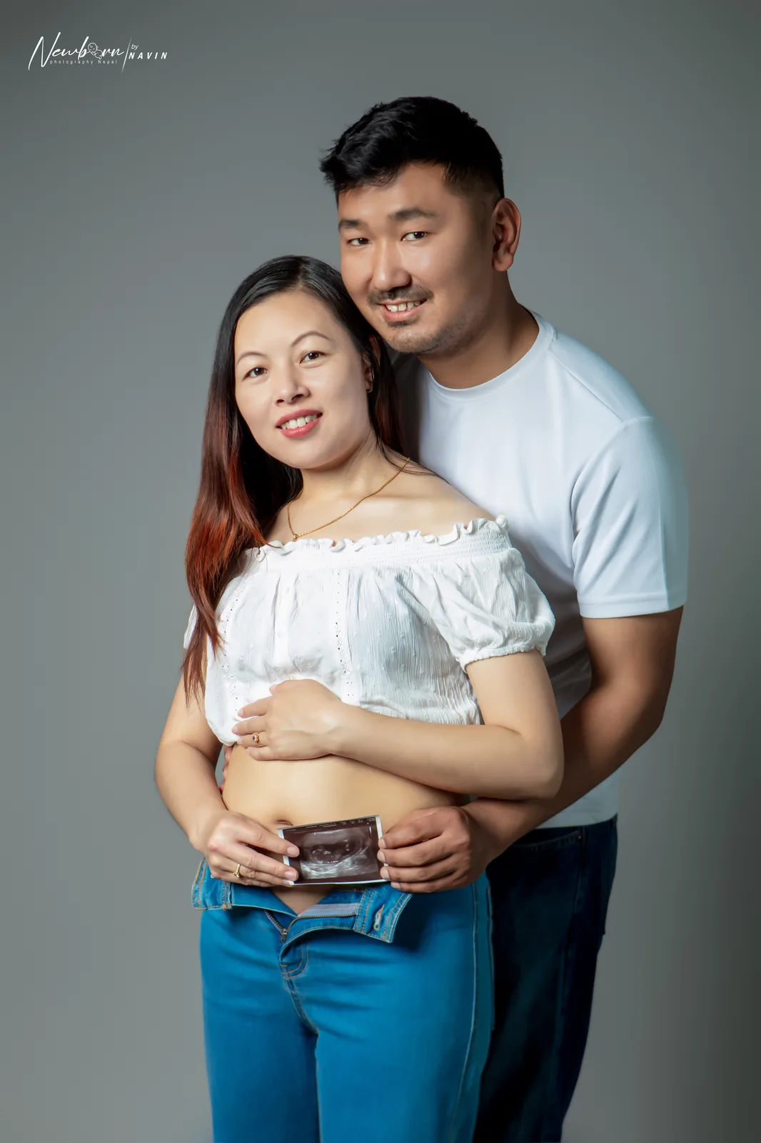 Expecting parents standing together, the father's arms around the mother, both hands holding an ultrasound photo against her bare bump.