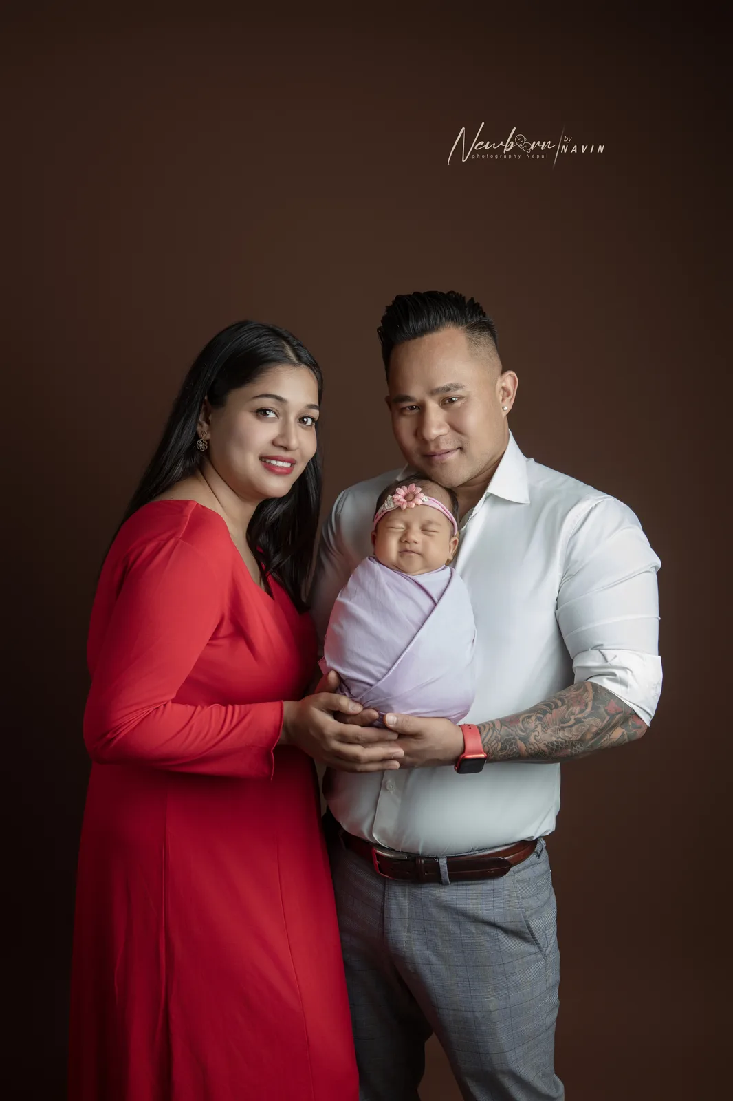Parents holding their newborn baby girl, wrapped in lavender with a flower headband, photographed against a dark brown backdrop.