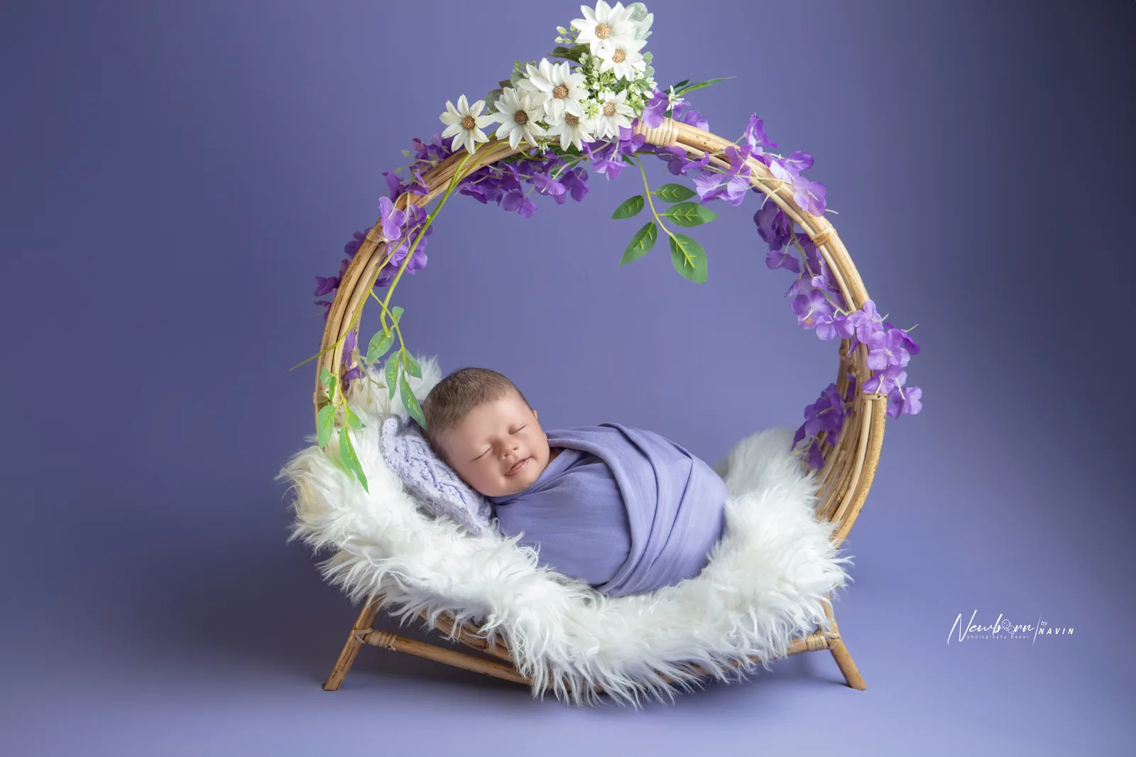 Newborn baby wrapped in a lavender knit wrap, sleeping inside a round rattan wreath decorated with purple and white flowers.