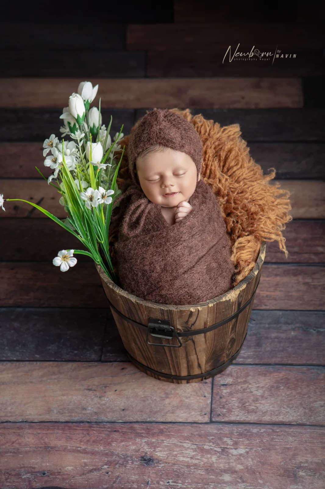 Newborn baby wrapped in a brown knit wrap and bonnet, sleeping in a wooden bucket prop styled with white flowers against a rustic wood-plank backdrop.