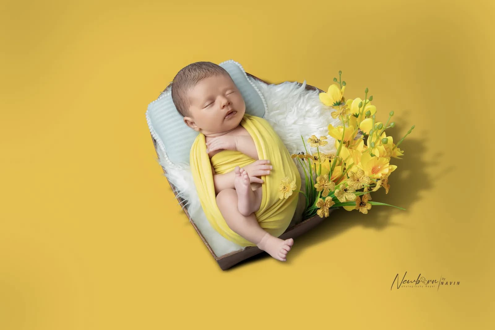 Newborn baby wrapped in a yellow wrap, sleeping on a wooden tray styled with yellow flowers against a warm yellow backdrop.