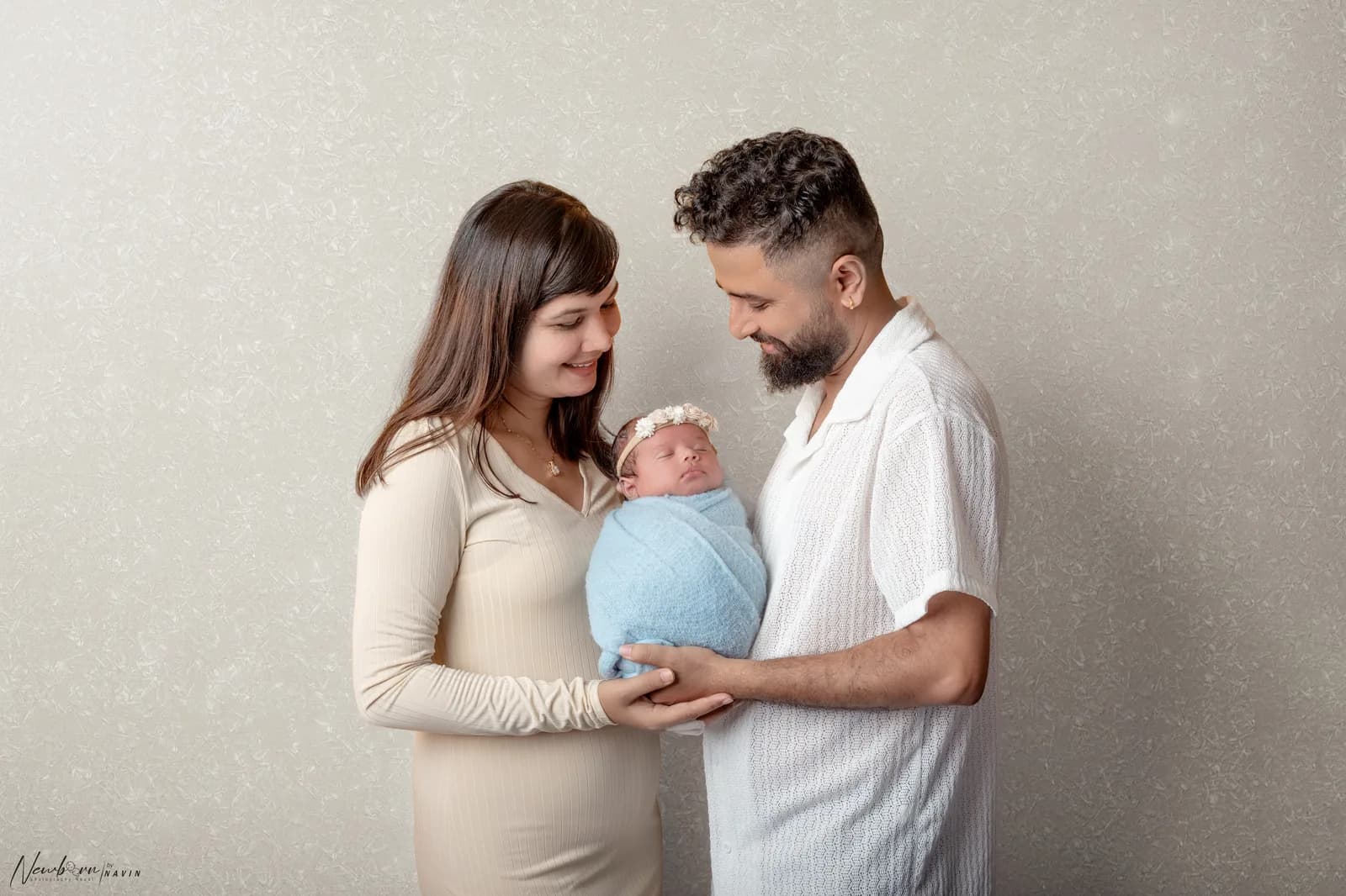 Mother and father smiling down at their newborn baby, wrapped in blue and wearing a floral headband, held between them.