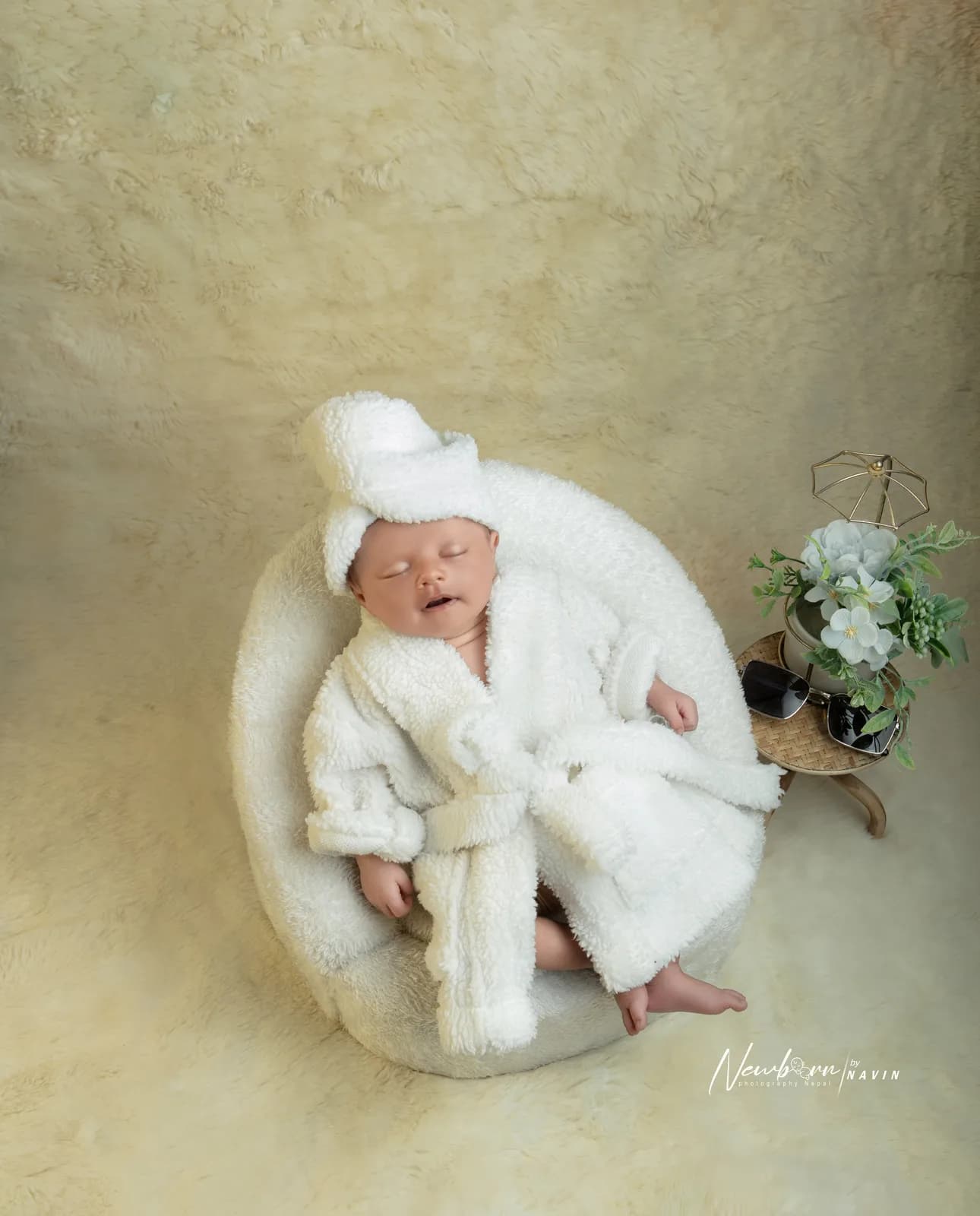 Newborn baby dressed in a miniature white spa robe and towel turban, sleeping in a round chair beside sunglasses and a small flower arrangement.