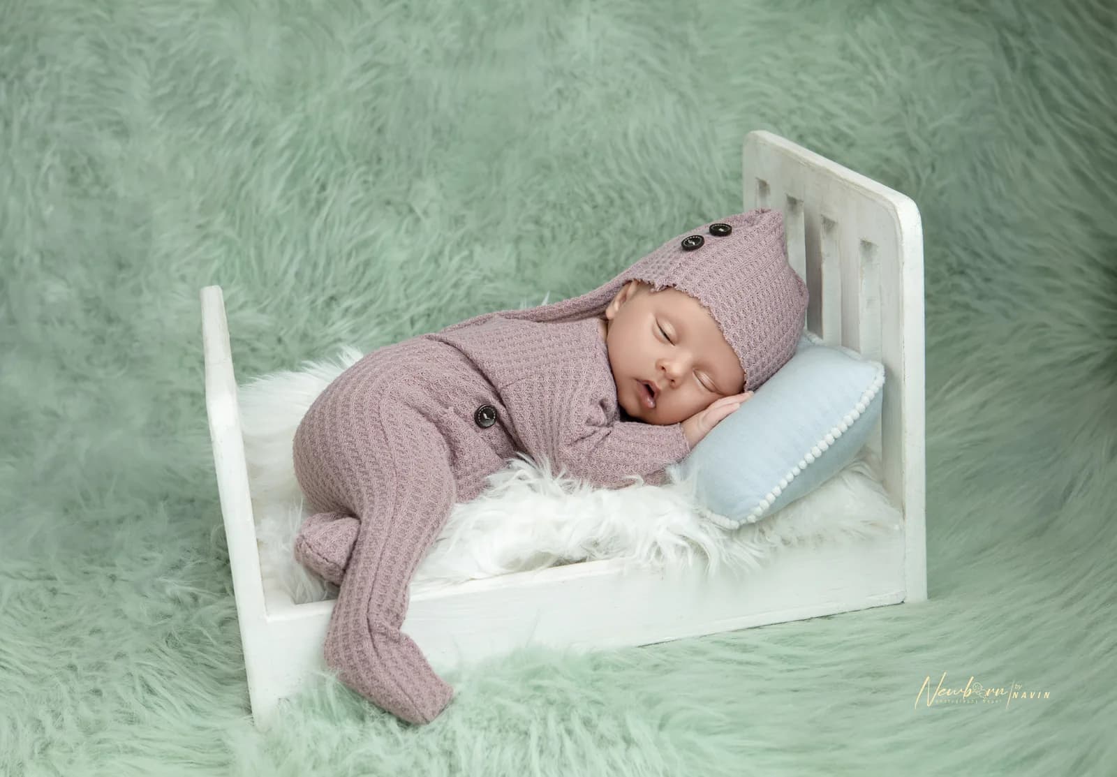 Newborn baby wrapped in a dusty pink knit outfit and bonnet, sleeping on a miniature white wooden bed with a blue pillow, against a green faux fur backdrop.