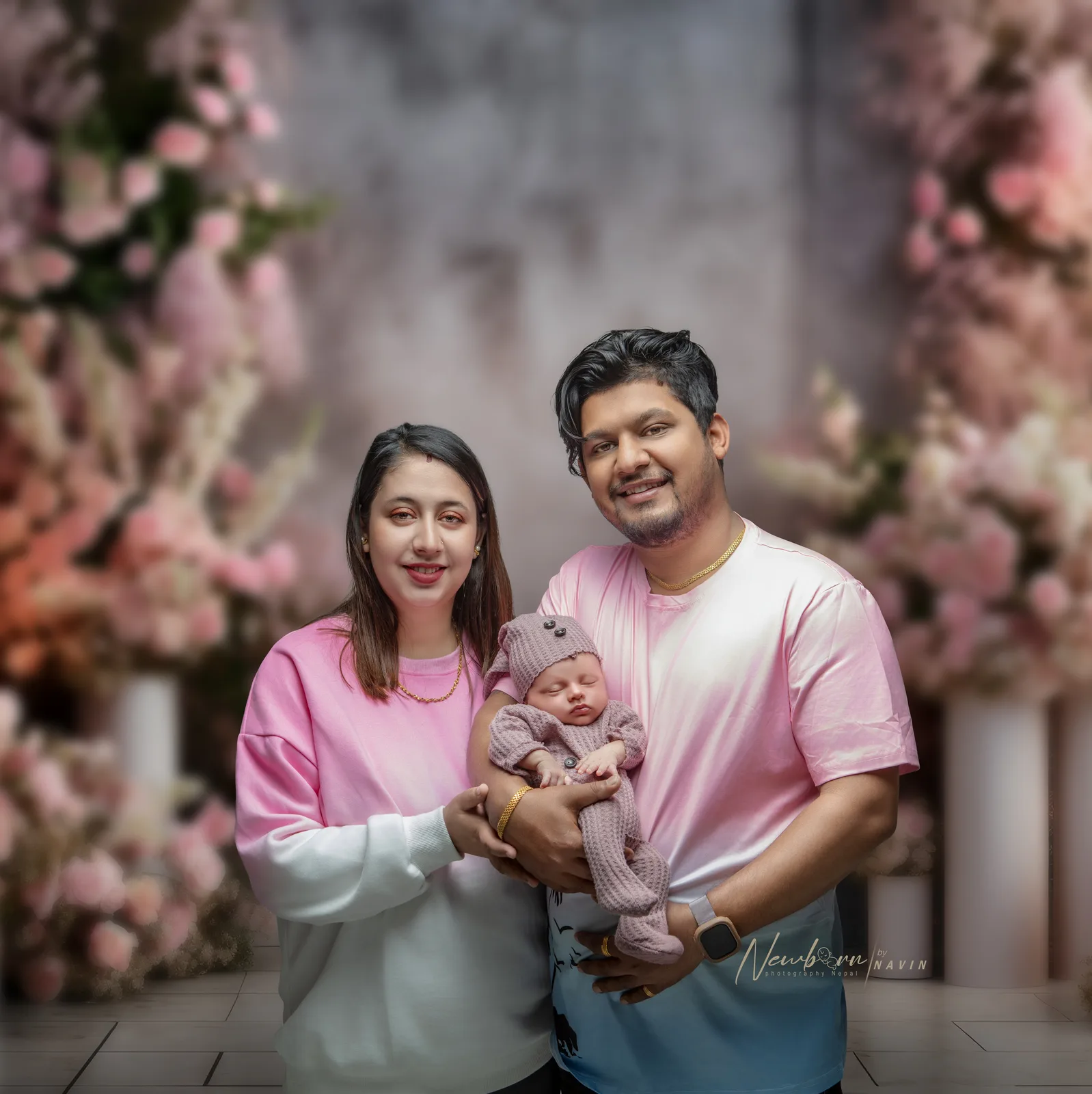Father with a tattooed arm smiling and holding his newborn baby beside a window with sheer curtains and a floral wall mural.