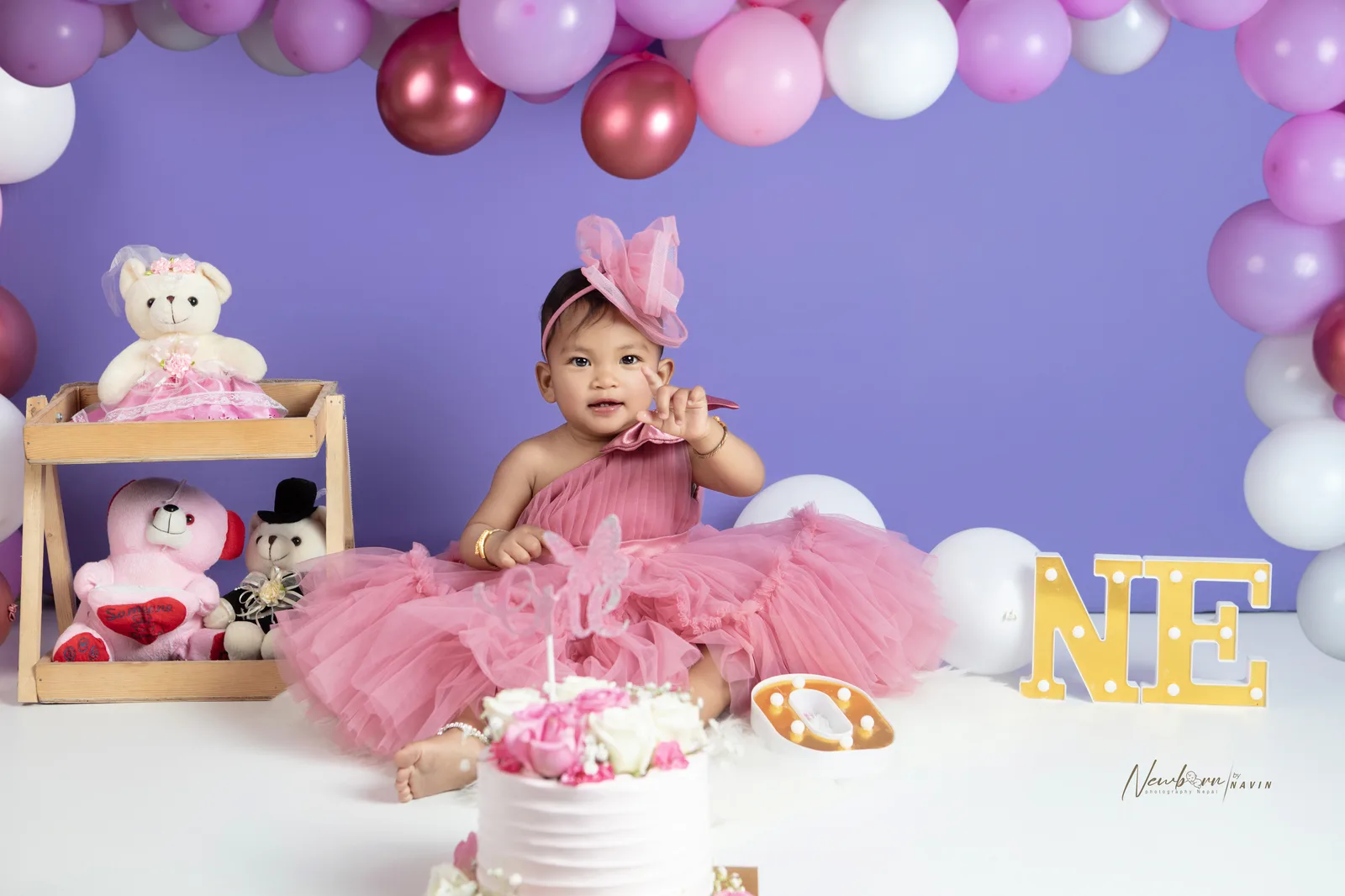 One-year-old girl in a pink tulle dress and fascinator sitting in front of a white cake decorated with roses, with a purple balloon garland and teddy bears in the background.