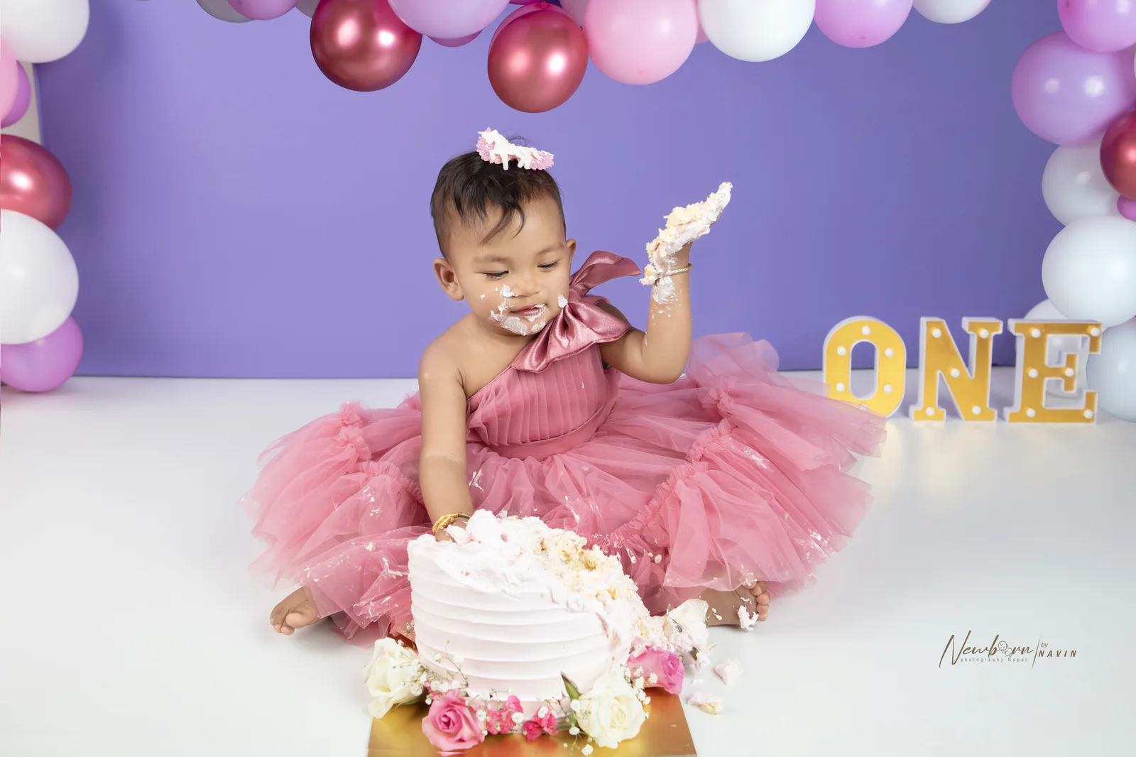 One-year-old girl with cake frosting on her face and her hand raised showing more frosting, sitting beside a smashed cake with an illuminated "ONE" sign behind her.