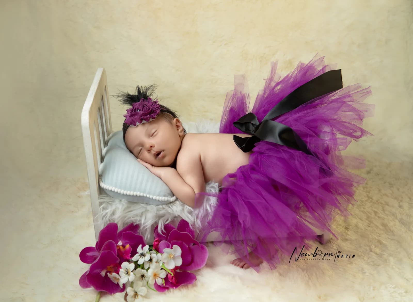 Newborn baby girl sleeping on a miniature wooden bed prop, wearing a purple tulle skirt and a flower headband, with purple and white flowers in the foreground.